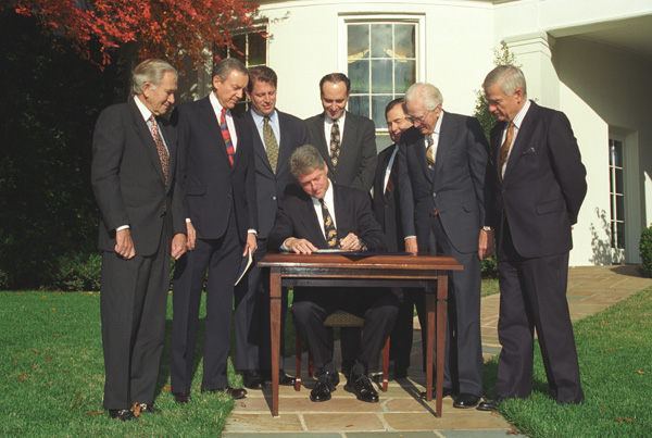 President Clinton sitting at a desk outside surrounded by men in suits signing RFRA into law
