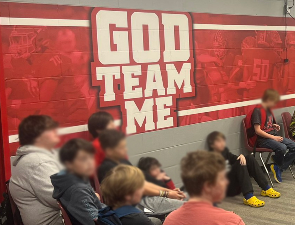 A group of school kids with blurred faces sit underneath a school spirit sign that reads "GOD Team Me"