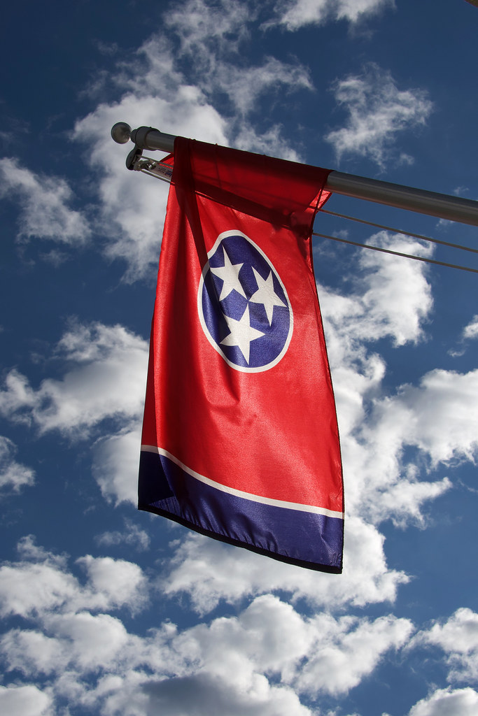 A shot of the Tennessee state flag, a red flag with a blue stripe on one side, in the center is a blue circle with three white stars. The background is a bright blue sky with cumulus clouds.